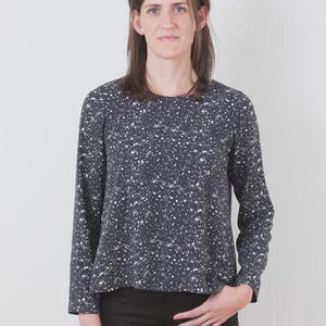 May include: A long-sleeved, dark gray top with a speckled white pattern. The top has a round neckline and is paired with black pants. The model is standing in front of a white background.