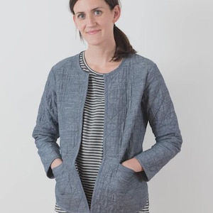 May include: A woman wearing a blue quilted jacket with a round neckline and two front pockets. Underneath, she wears a striped black and white shirt. The jacket is open, revealing the shirt. She is also wearing blue jeans.