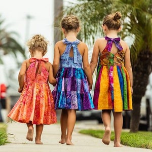 May include: Three young girls walking away, holding hands, wearing colorful halter dresses. The dresses feature vibrant patterns and bow details at the back. The dresses are in shades of orange, blue, purple, yellow, and green.