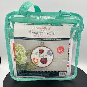 May include: A clear, turquoise-handled tote bag containing a Kimberbell Punch Needle Embroidery kit. The kit includes a round embroidery hoop with a ladybug and flower design. The text on the kit reads "BUGS & BLOOMS KIT" and "Punch Needle Embroidery".