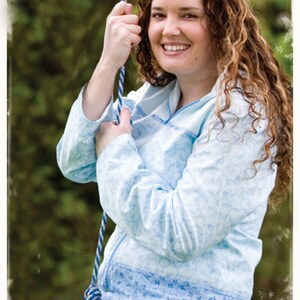 May include: A woman with long curly brown hair is wearing a light blue jacket with a white floral pattern. She is holding a blue and white rope in her hands. The woman is smiling and looking at the camera.