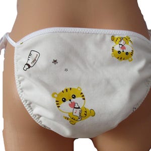 May include: White fabric diaper cover featuring a repeating pattern of yellow cartoon tigers holding baby bottles. The garment has white trim along the edges and white side ties for closure. Small black star icons are scattered across the fabric.