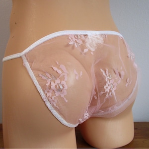 May include: Sheer light pink underwear featuring a delicate white floral lace pattern. The garment is constructed from a translucent mesh fabric and is finished with a thin white elastic trim along the waistband and leg openings.
