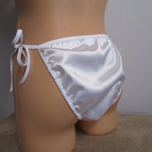 May include: A pair of white satin bikini-style underwear featuring a smooth, glossy finish and thin side ties that form bows. The garment has a high-cut leg design with white trim along the edges and a gathered detail along the side seams.