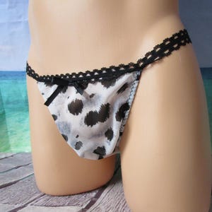 May include: A white thong featuring a black animal print pattern, accented with a delicate black lace trim along the waistband and a small black ribbon bow centered at the front. The garment is constructed from a lightweight, smooth fabric.