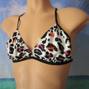 May include: A triangle bikini top featuring a white base with a black leopard print pattern and subtle accents of orange and pink. The garment is designed with thin black straps and black trim along the edges of the cups and the under-bust band.