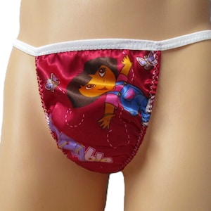 May include: A red satin thong featuring a graphic print of the cartoon character Dora the Explorer, blue overalls, and purple butterflies. The garment is finished with a white elastic waistband and white trim along the edges of the fabric.