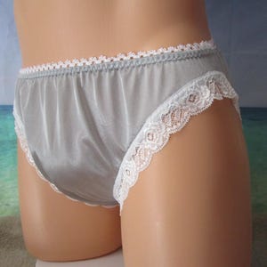 May include: A pair of silver-grey briefs featuring a smooth, satin-like finish. The garment is accented with a delicate white scalloped lace trim along the waistband and the leg openings, providing a decorative edge to the solid grey fabric.