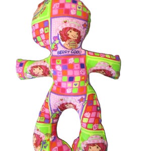 May include: A colorful, handmade doll featuring a patchwork design with images of a cartoon character. The doll has a head, arms, and legs, and the text "BERRY COOL!" is visible. The doll is primarily pink, green, and orange.