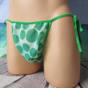 May include: A sheer white bikini bottom featuring a green polka dot pattern. The garment includes thin green side ties for an adjustable fit and is finished with green trim along the edges of the fabric, creating a simple and functional design.