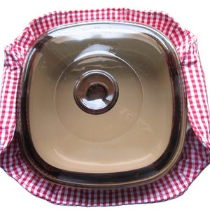 Könnte beinhalten: Ein quadratischer, klarer Glas-Auflaufform mit Deckel, der in einer rot-weiß karierten Stoffhülle liegt. Die Form hat einen silbernen Rand und einen zentralen Knopf auf dem Deckel. Die Stoffhülle sorgt für einen dekorativen Touch.
