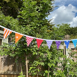 May include: A rainbow-coloured bunting with different patterns on each flag. The bunting is strung between two trees in a garden.