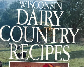 Könnte beinhalten: Ein spiralgebundenes Kochbuch mit dem Titel Wisconsin Dairy Country Recipes: A Collection from America's Dairyland. Das Cover zeigt eine grüne Weide mit schwarz-weißen Kühen und ein zentrales Bild verschiedener Käsesorten, Milchflaschen und Sahne.