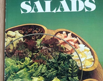 Könnte beinhalten: Ein Kochbuch mit weichem Einband mit dem Titel Betty Crocker's Salads und grünem Cover. Die Vorderseite zeigt eine große Glasschüssel mit gemischtem grünem Salat, umgeben von Holzschalen mit Oliven, Schinken, Eiern, Speck und Gurken.