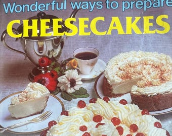 Könnte beinhalten: Ein Kochbuch mit dem Titel Wonderful ways to prepare CHEESECAKES, das einen großen Käsekuchen mit Schlagsahne und roten Kirschen auf einem roten Glasteller, ein Stück auf einem kleinen Teller und einen ganzen Käsekuchen zeigt.