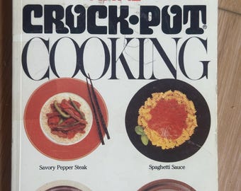 Könnte beinhalten: Ein weißes Taschenbuch-Kochbuch mit dem Titel RIVAL CROCK-POT COOKING. Das Cover zeigt vier kreisförmige Bilder von Speisen: Savory Pepper Steak, Spaghetti Sauce, Swiss Steak und Cheesecake. Der Text lautet: Mehr als 300 Rezepte für den Slow Cooker.