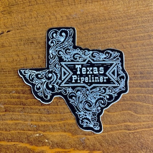 May include: A black and white sticker in the shape of Texas. The sticker features ornate scrollwork and the words "Texas Pipeliner" in a banner. The sticker is on a wooden surface.