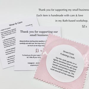 May include: A close-up of a pink polishing cloth for silver and gold jewelry, with a white label. The label provides instructions for use. There are also care instruction cards and a thank you card.
