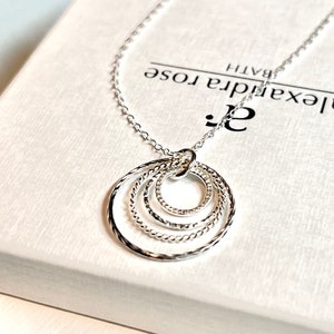 May include: A silver necklace with a pendant featuring four interlocking circles. The circles are textured and have a delicate design. The necklace is displayed on a white box with the text "Alexandra Rose Bath" printed on it.