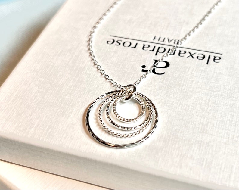 May include: A silver necklace with a pendant featuring four interlocking circles. The circles are textured and have a delicate design. The necklace is displayed on a white box with the text "Alexandra Rose Bath" printed on it.