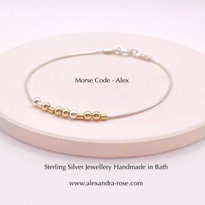 May include: A silver bracelet with a delicate chain and a row of alternating gold and silver beads. The bracelet is on a white surface and the text "Morse Code - Alex" is visible above the bracelet.