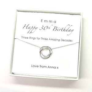 May include: A white gift box with a silver necklace inside. The necklace has three interlocking rings. The box has the text "Emma Happy 30th Birthday Three Rings for Three Amazing Decades Love from Anna x" printed on it.