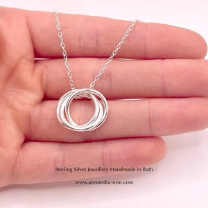 May include: A silver necklace with a pendant made of three interlocking rings. The necklace is on a hand, and the text "Sterling Silver Jewellery Handmade in Bath www.alexandra-rose.com" is below the necklace.