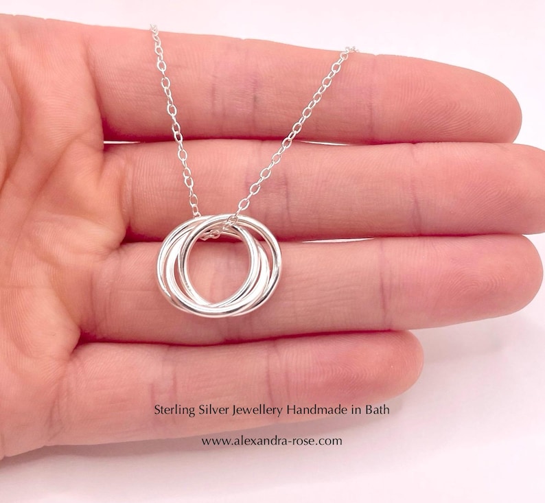 May include: A silver necklace with a pendant made of three interlocking rings. The necklace is on a hand, and the text "Sterling Silver Jewellery Handmade in Bath www.alexandra-rose.com" is below the necklace.
