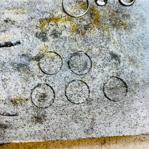 May include: A close-up of a gray, textured surface with six circular indentations. The surface appears to be a workbench or a table used for crafting or jewelry making.