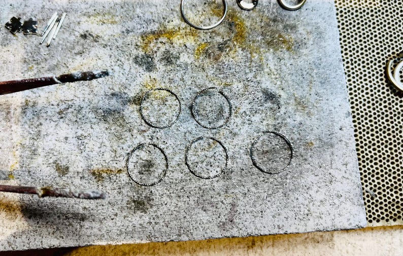May include: A close-up of a gray, textured surface with six circular indentations. The surface appears to be a workbench or a table used for crafting or jewelry making.
