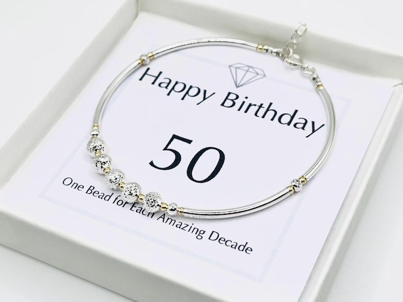 May include: Silver-tone bracelet with gold accents and decorative beads. The bracelet is presented on a white card with the text "Happy Birthday 50" and "One Bead for Each Amazing Decade."