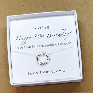 May include: A white gift box with a silver necklace inside. The necklace has three interlocking rings. The box has a message that reads "Katie Happy 30th Birthday! Three Rings for Three Amazing Decades Love from Lucy x".
