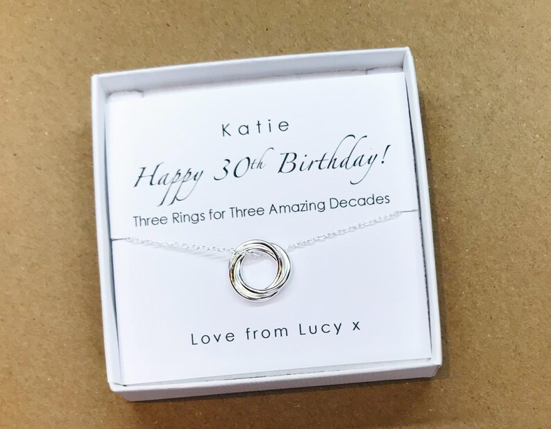 May include: A white gift box with a silver necklace inside. The necklace has three interlocking rings. The box has a message that reads "Katie Happy 30th Birthday! Three Rings for Three Amazing Decades Love from Lucy x".