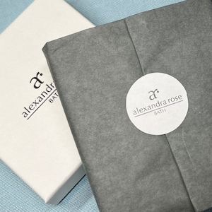 May include: A white box and a wrapped package are displayed on a light blue surface. The box has the text "alexandra rose BATH" printed in black. The wrapped package is gray and has a white circular sticker with the same text.