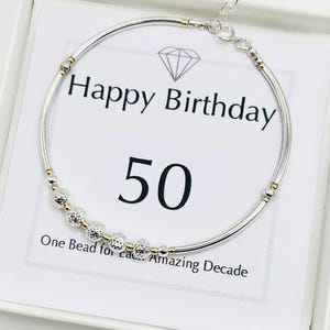 May include: A silver bracelet with gold accents and a series of textured beads. The bracelet is displayed on a card that reads "Happy Birthday 50" with the text "One Bead for Each Amazing Decade".