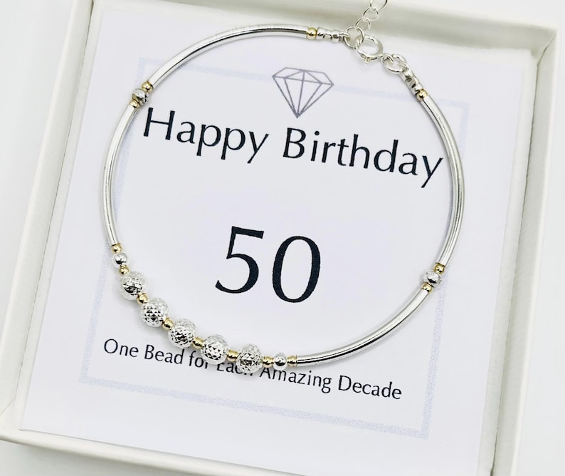 May include: A silver bracelet with gold accents and a series of textured beads. The bracelet is displayed on a card that reads "Happy Birthday 50" with the text "One Bead for Each Amazing Decade".