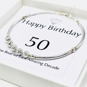 May include: A silver bracelet with decorative beads and gold accents, presented on a white card. The card reads "Happy Birthday 50" with the text "One Bead for Each Amazing Decade."