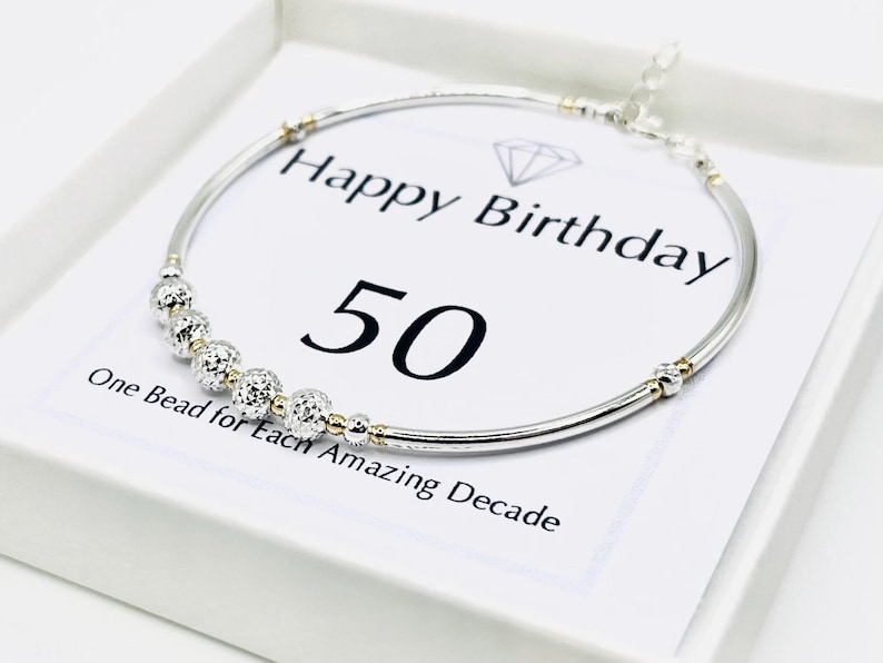 May include: A silver bracelet with decorative beads and gold accents, presented on a white card. The card reads "Happy Birthday 50" with the text "One Bead for Each Amazing Decade."