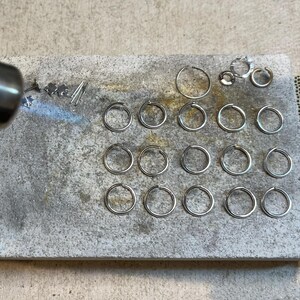 May include: A close-up of a metal work surface with a torch and a variety of silver rings. The rings are arranged in rows and are being heated with a torch.