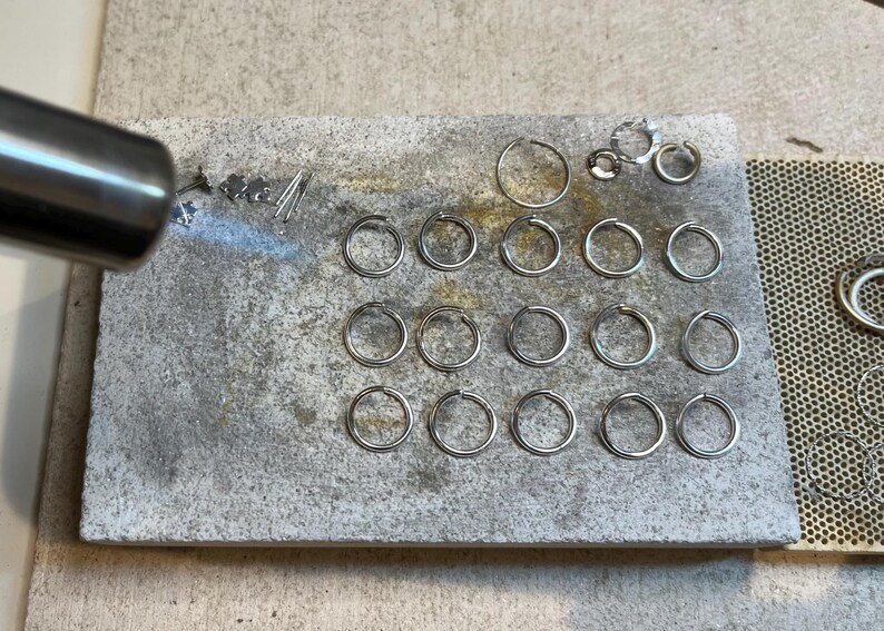 May include: A close-up of a metal work surface with a torch and a variety of silver rings. The rings are arranged in rows and are being heated with a torch.