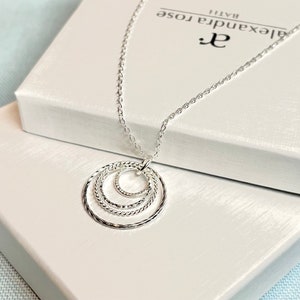 May include: Silver necklace with a pendant featuring three interlocking circles. The circles are textured and have a delicate, feminine design. The necklace is displayed on a white box with the brand name "Alexandra Rose" printed on it.