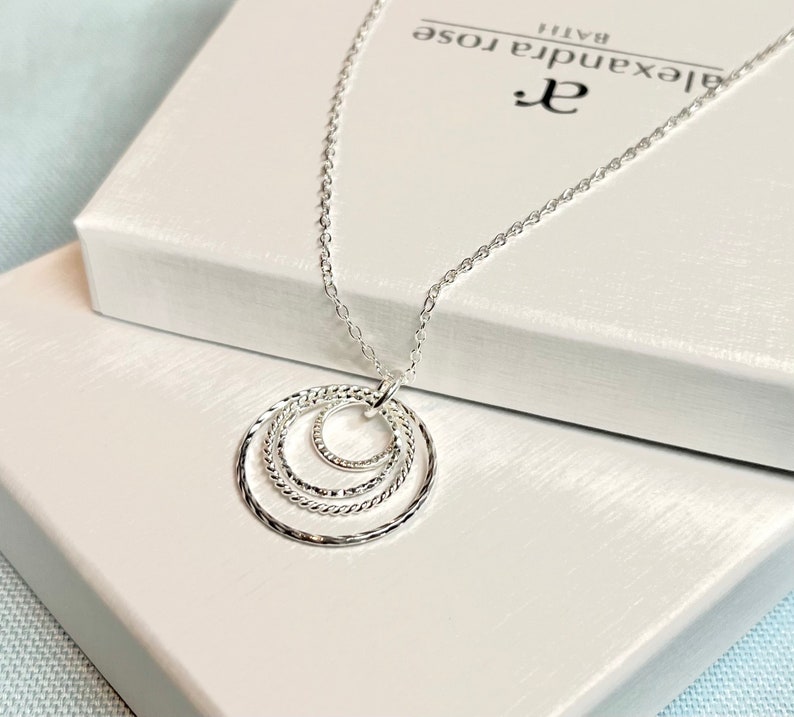 May include: Silver necklace with a pendant featuring three interlocking circles. The circles are textured and have a delicate, feminine design. The necklace is displayed on a white box with the brand name "Alexandra Rose" printed on it.