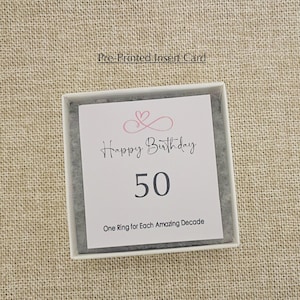 May include: A white card with a pink heart design and the text "Happy Birthday 50 One Ring for Each Amazing Decade".