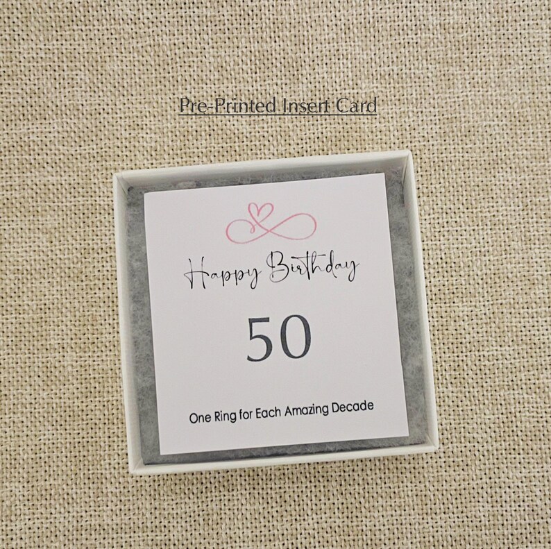 May include: A white card with a pink heart design and the text "Happy Birthday 50 One Ring for Each Amazing Decade".