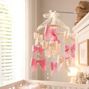 May include: A hanging nursery mobile featuring multiple fabric bows in shades of pink and cream, suspended from a circular wooden frame by thin strings adorned with small white beads, topped with a sheer cream-colored ribbon bow.