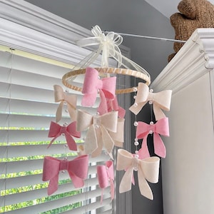 May include: A hanging mobile featuring multiple felt bows in shades of soft pink and cream, suspended from a circular wooden hoop by thin strings accented with small white faux pearls. A sheer white ribbon bow is tied at the top center point.