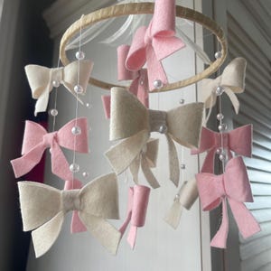 May include: A hanging mobile featuring a circular wooden hoop adorned with multiple felt bows in shades of soft pink and cream. The bows are suspended by thin white strings interspersed with small white beads, and a white ribbon bow sits at the top.