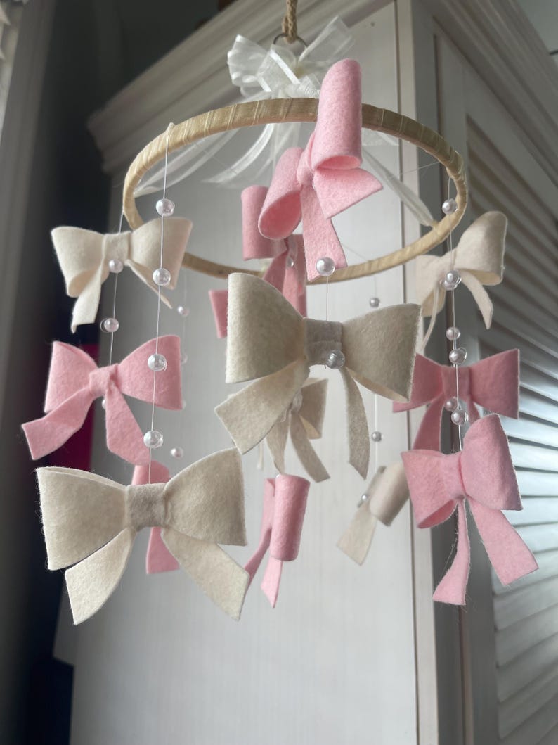 May include: A hanging mobile featuring a circular wooden hoop adorned with multiple felt bows in shades of soft pink and cream. The bows are suspended by thin white strings interspersed with small white beads, and a white ribbon bow sits at the top.