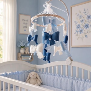 May include: A nursery mobile featuring multiple fabric bows in shades of navy blue, light blue, and white, suspended from a circular frame with white ribbons and small white beads. The mobile hangs from a white metal arm attached to a white crib.