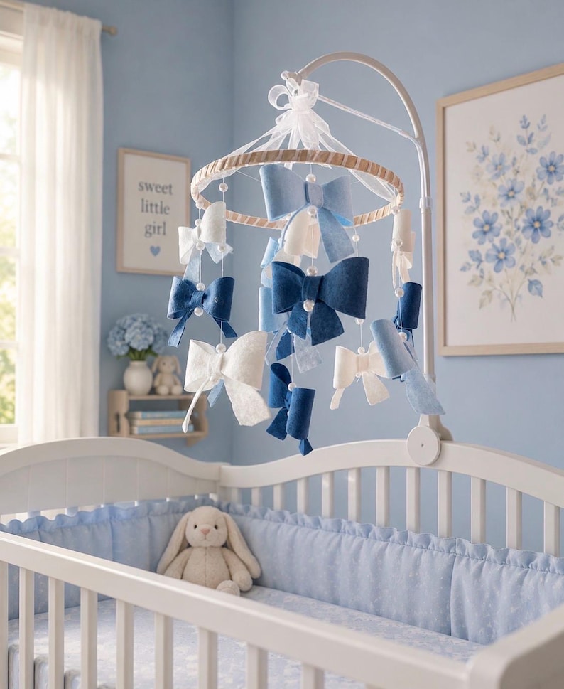 May include: A nursery mobile featuring multiple fabric bows in shades of navy blue, light blue, and white, suspended from a circular frame with white ribbons and small white beads. The mobile hangs from a white metal arm attached to a white crib.